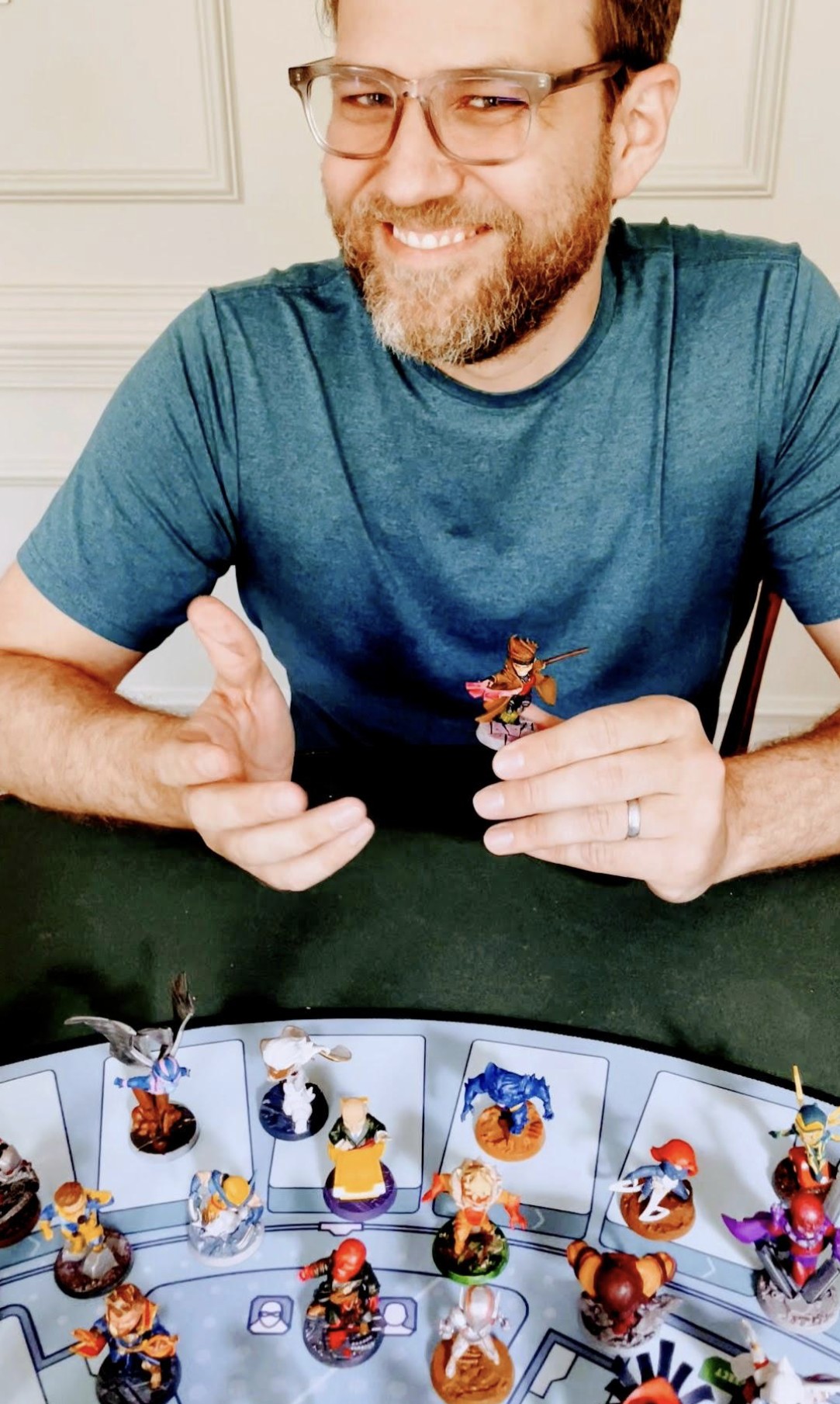 Jim smiling at the table, holding up a painted mini above a Marvel United board full of painted heroes
