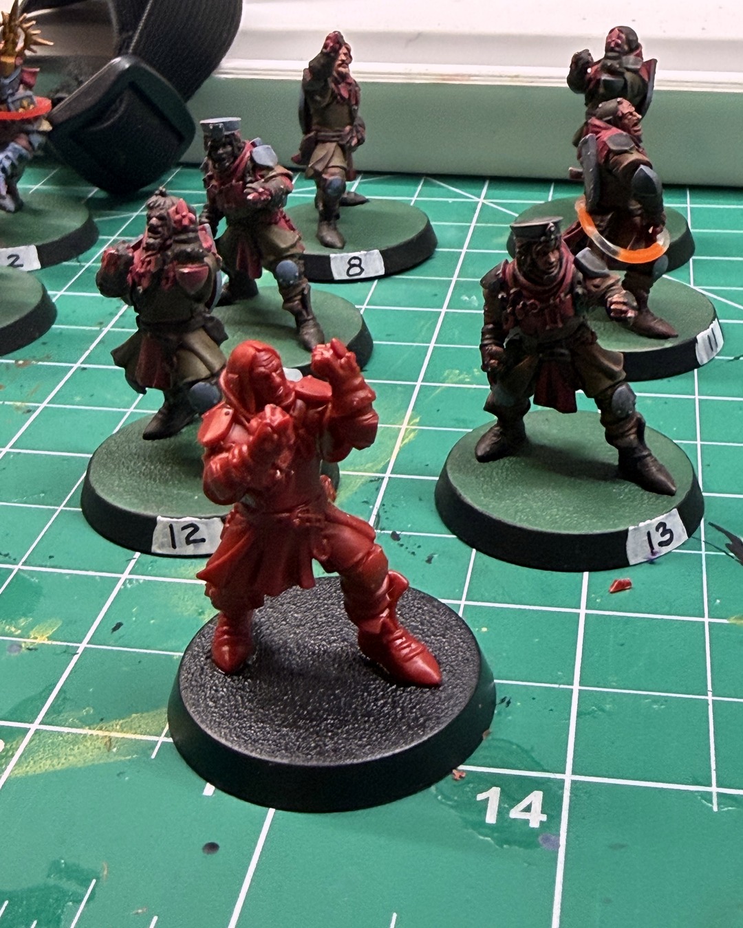 A newly built, unpainted red plastic squire on a black base standing in front of painted squires numbered 12 and 13