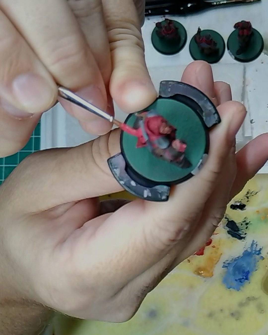Overhead close-up of a brush laying red onto a squire held on a painting handle, with a wet palette and more squires in the background