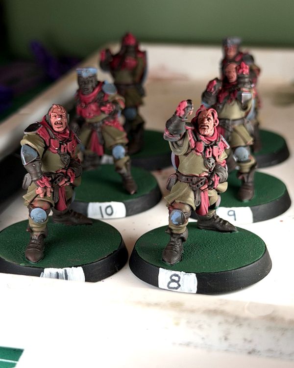 Holy Hand Grenadiers squires numbered 8 and 11 in focus, olive tunics with red scarves and slate blue knee pads, on green bases with white number labels, more squires blurred behind