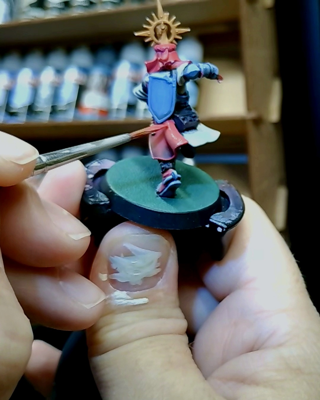 A knight with a gold sunburst halo and blue shield held on a painting handle while a brush highlights his red cape, paint racks blurred behind