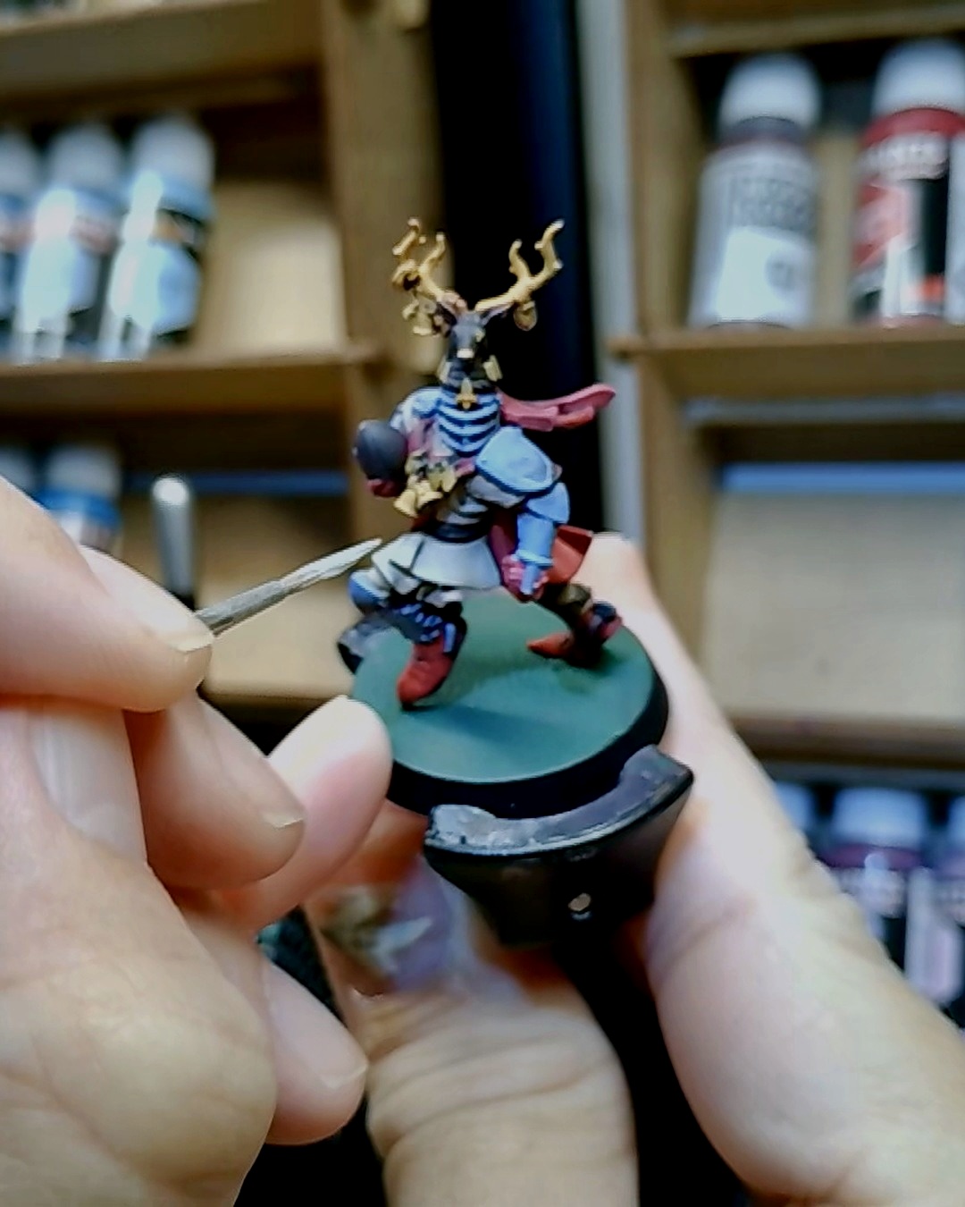 The stag-helm knight held on a painting handle with a brush working highlights onto the armor, Vallejo paint bottles on the rack behind