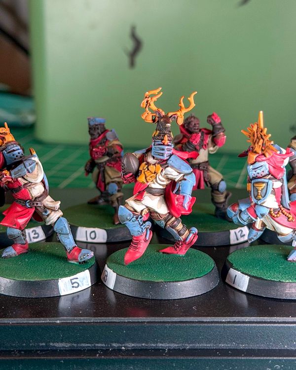 Holy Hand Grenadiers knight number 8 in a stag-antler helm carrying the ball, slate blue armor and red cape, in focus between knights 5 and 1 on green bases with white rim numbers