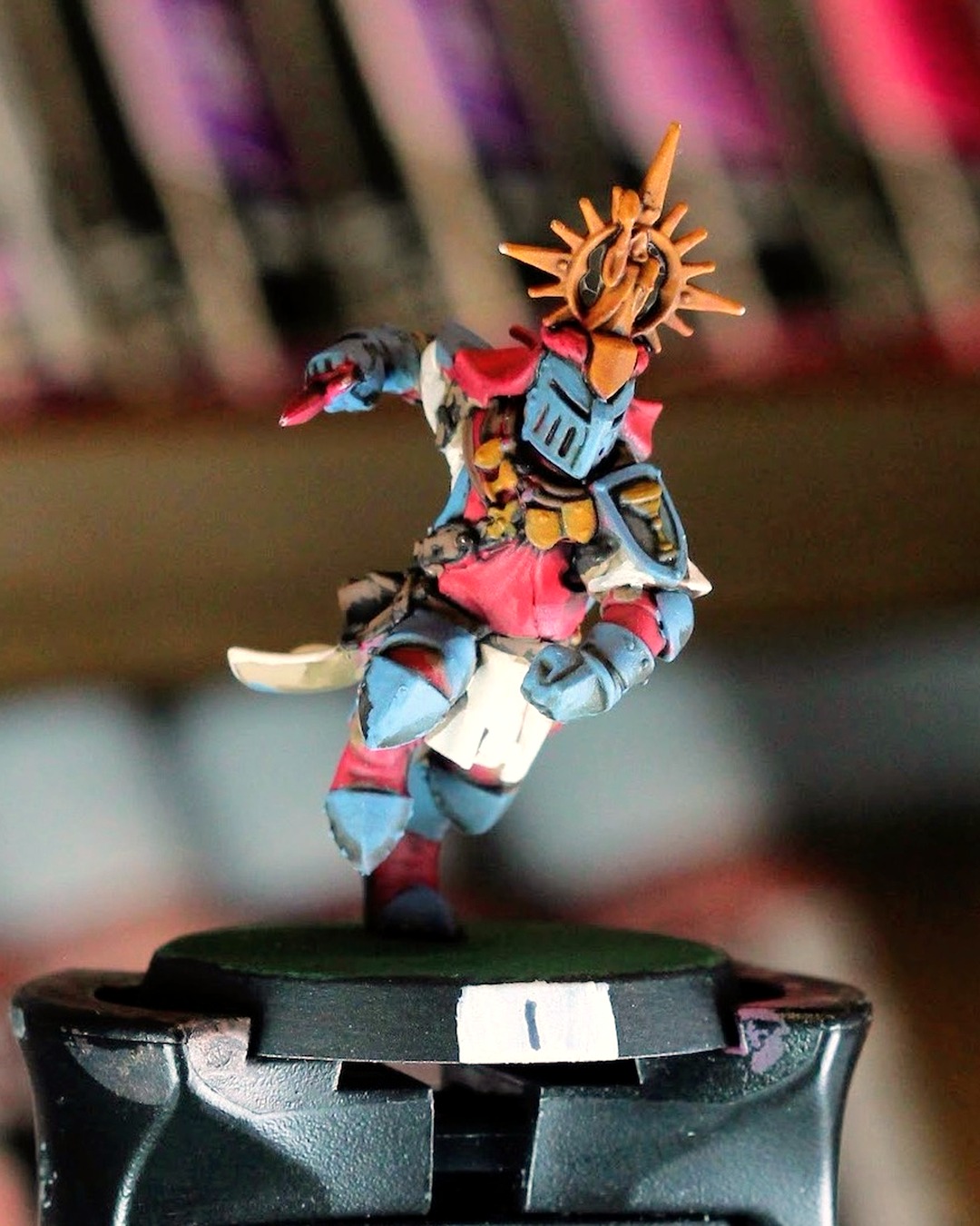 Holy Hand Grenadiers knight number 1 on a painting handle, gold sunburst halo, slate blue armor, red tabard and white tassets, paint racks blurred behind