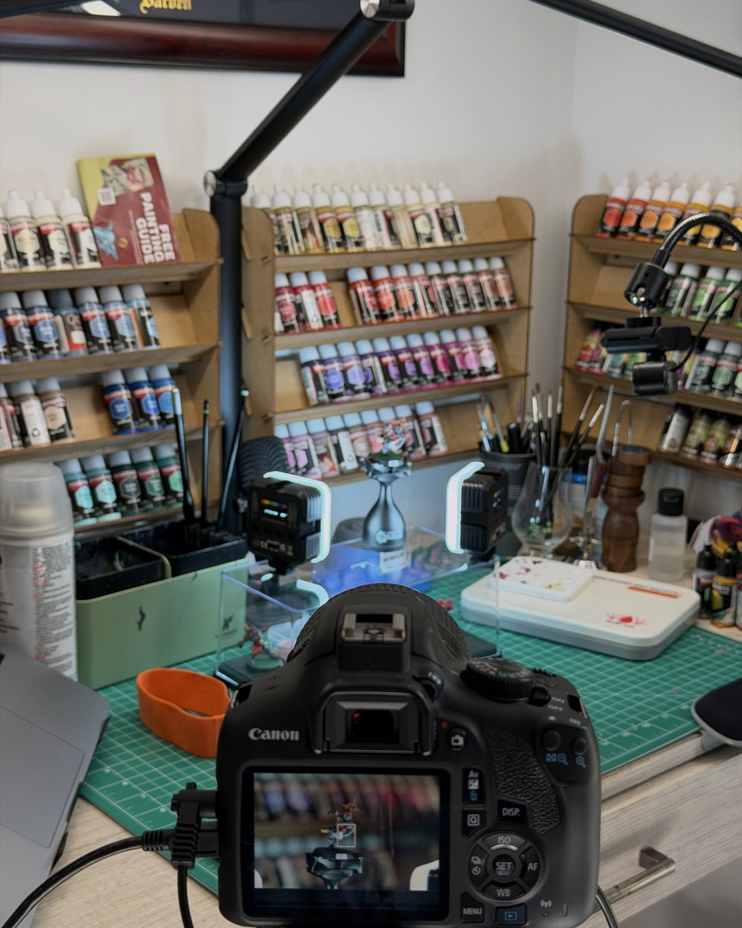 Over-the-shoulder view of the Canon Rebel at the painting desk, a Blood Bowl knight on a painting handle between two small LED lights, wall paint racks behind and the mini on the camera's rear screen