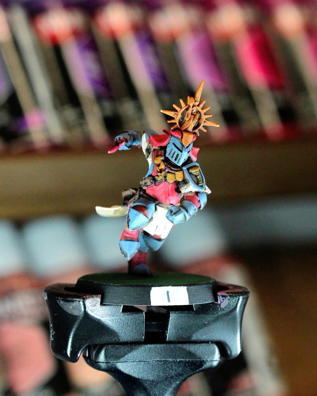 The resulting shot: Holy Hand Grenadiers knight number 1 on a painting handle, gold sunburst halo, slate blue armor and red tabard, paint racks melted into a soft blur behind