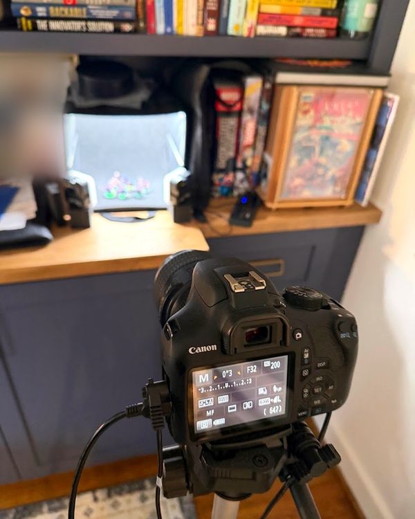 A Canon Rebel on a tripod, tethered by cable, aimed at a small glowing lightbox of painted minis on a wooden shelf; the rear screen shows manual mode, f/32, 0.3 seconds, ISO 200