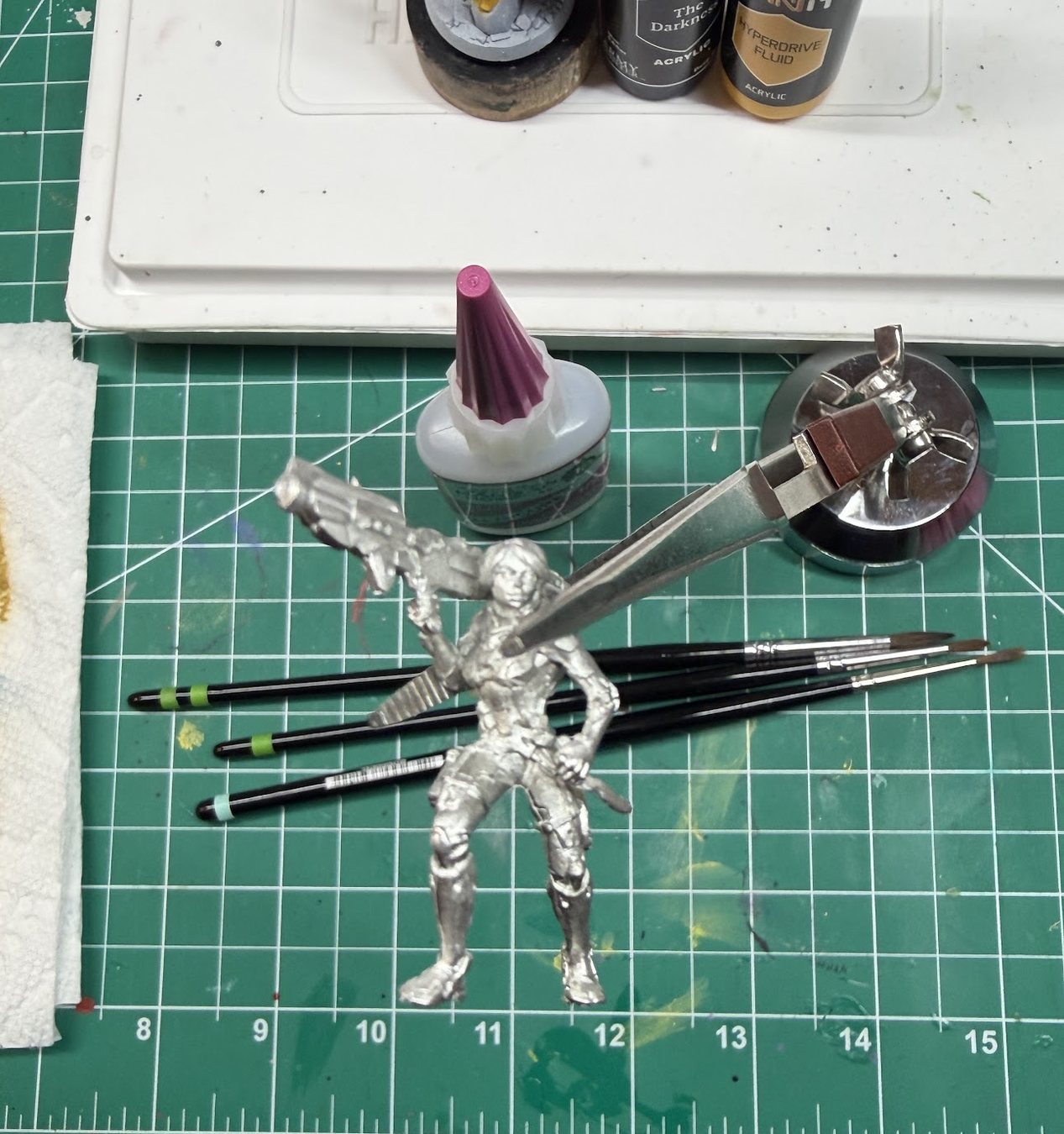 Three striped brushes on the cutting mat next to an unpainted metal mini held in a tweezer clamp